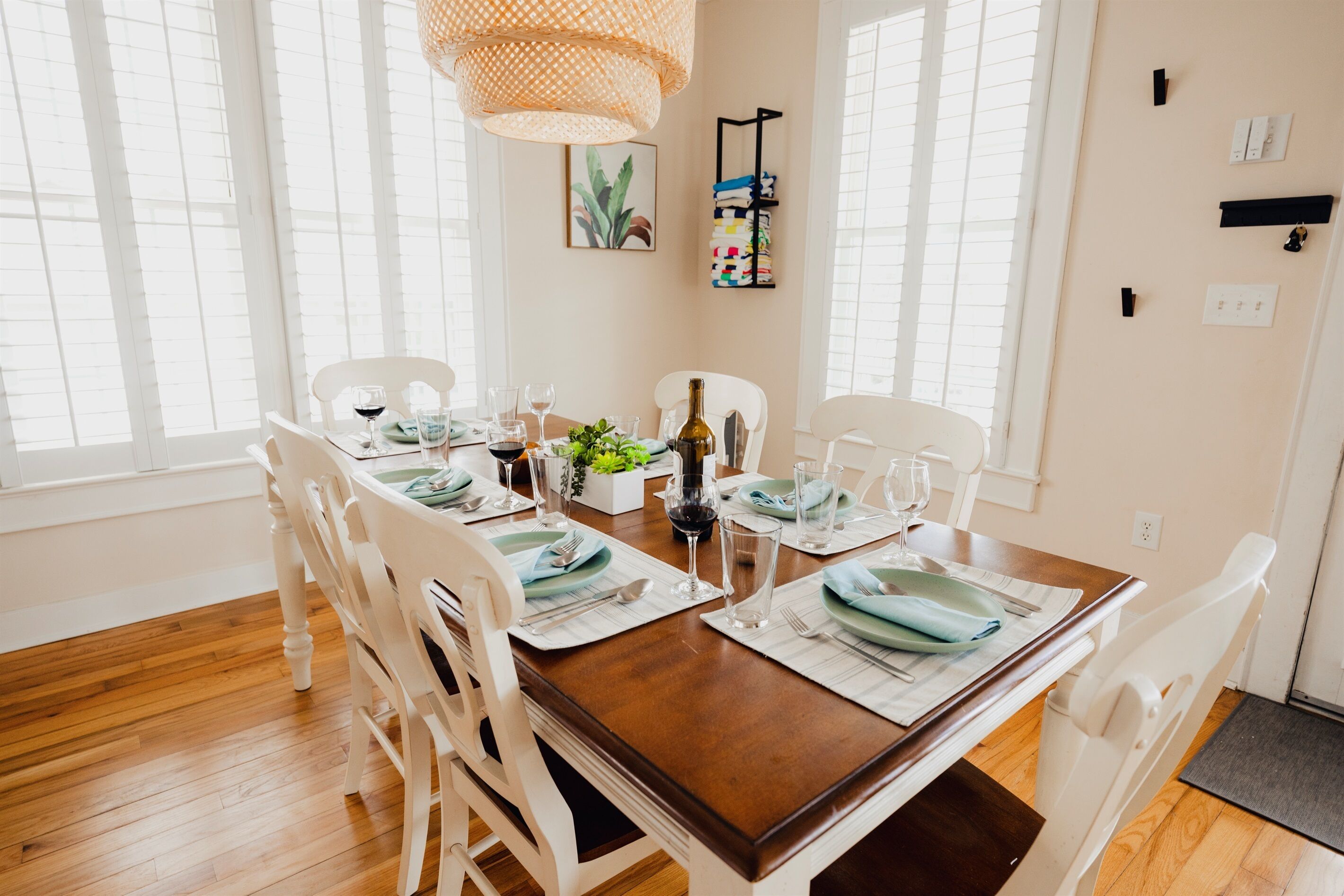 A dining area with a wooden table set for four, white chairs, a hanging light fixture, and a wall-mounted shelf with a plant.