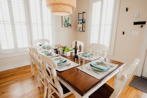 A dining area with a wooden table set for four, white chairs, a hanging light fixture, and a wall-mounted shelf with a plant.