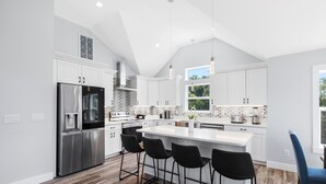 Une cuisine moderne avec des meubles blancs, un réfrigérateur en acier inoxydable, un grand îlot central doté de tabourets, et une vue sur l’extérieur par une fenêtre.