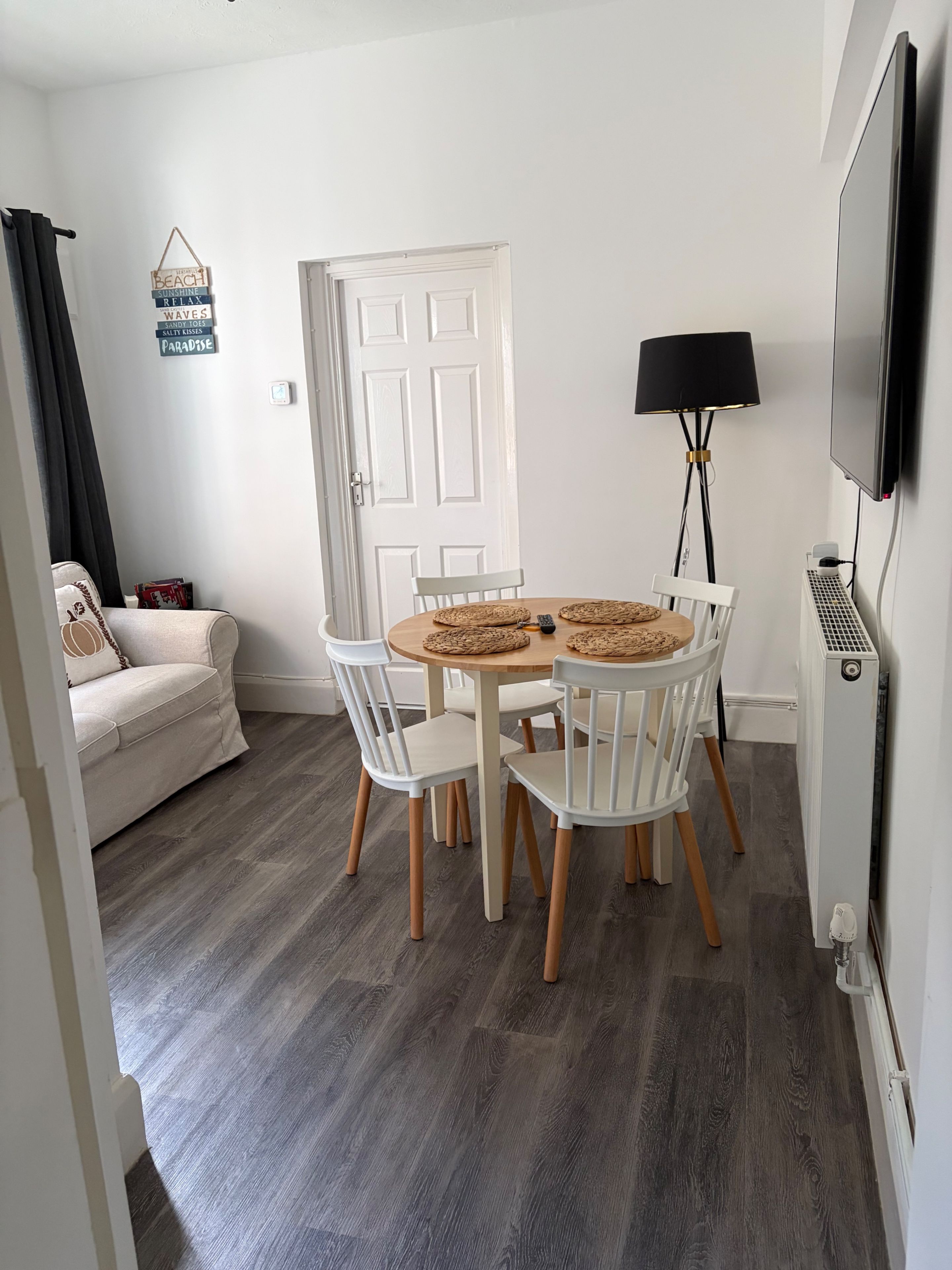 Une pièce avec une table ronde en bois, des chaises blanches, un canapé, une lampe et un téléviseur.
