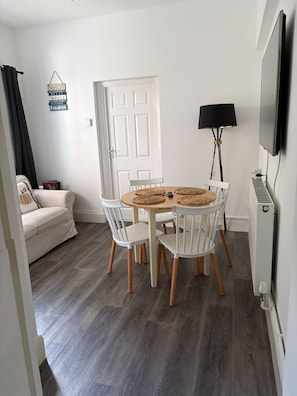 Una habitación con una mesa redonda de madera, sillas blancas, un sofá, una lámpara y un televisor.