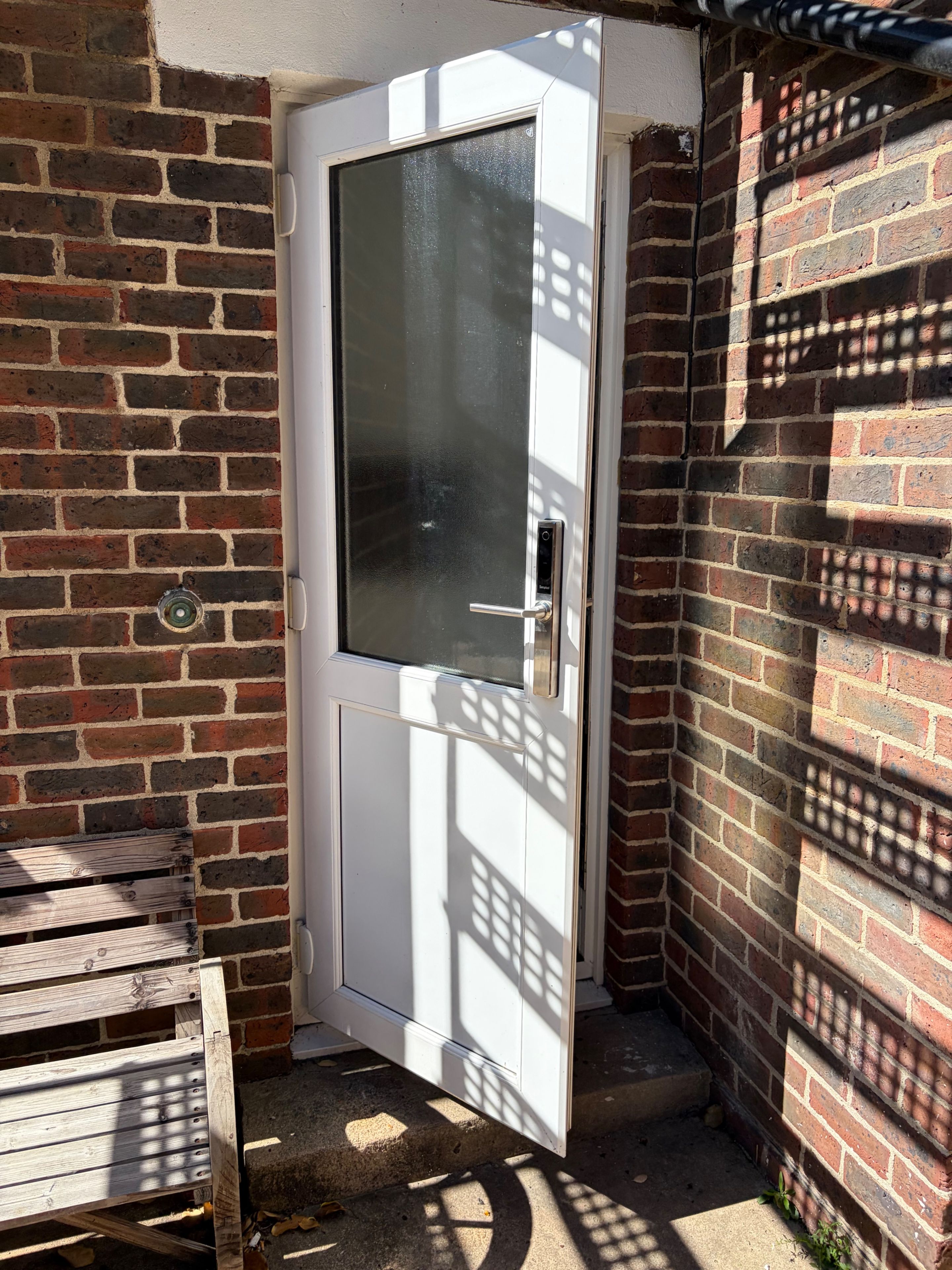 Eine weiße Tür mit Glasfenster, eingebettet in eine Hausecke mit Backsteinwand und einer Holzbank.