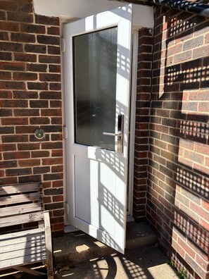 Una puerta blanca con un panel de vidrio, ubicada en una esquina junto a una pared de ladrillo y un banco de madera.