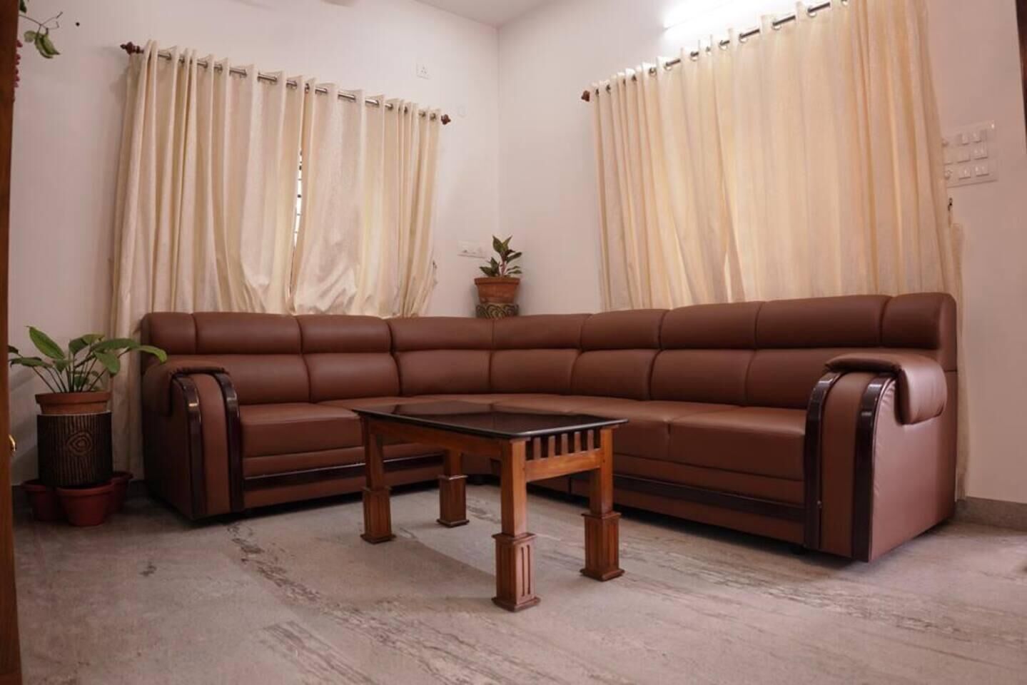 A living room with a brown leather sofa, a wooden coffee table, cream-colored curtains, and a potted plant.