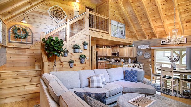 A cozy cabin interior with a wooden staircase, a living room with a sectional sofa, a dining area, and a kitchen.