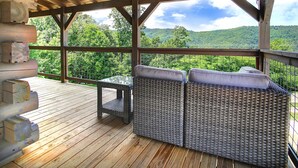 A covered wooden deck with wicker furniture and a glass table, overlooking a lush green landscape.