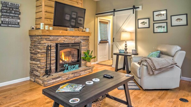 A cozy living room with a stone fireplace, a leather sofa, and a flat-screen TV mounted above it. - Country Cottage < 3 Mi to Dale Hollow Lake! (Byrdstown)