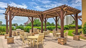 Un área de patio al aire libre con una pérgola, pilares de piedra, muebles de mimbre y vistas a un jardín ajardinado y edificios al fondo.