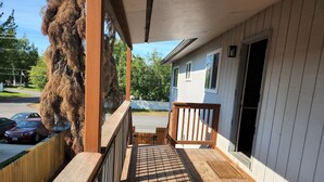 Une terrasse en bois avec une rambarde, un porche couvert et une vue sur les voitures garées et les arbres.
