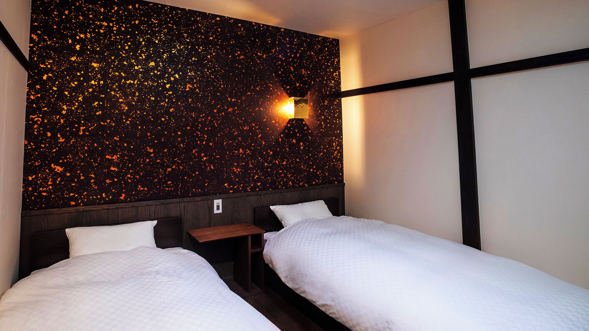 A hotel room with two beds, a nightstand, a wall-mounted light, and a textured wall.