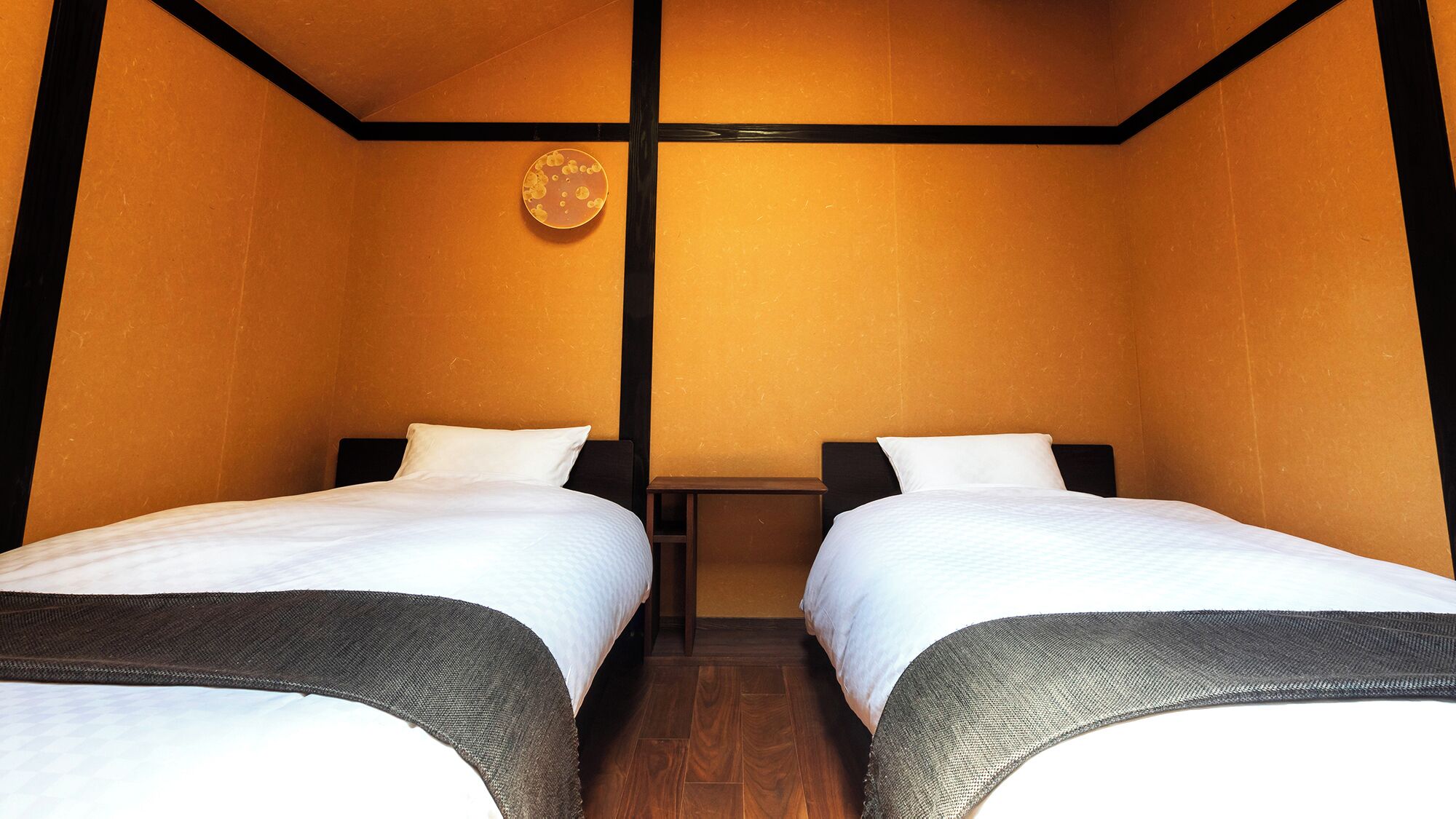 A room with two beds, wooden flooring, and a wall-mounted light fixture.