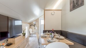 Un espace repas moderne avec une table en bois dressée pour quatre personnes, un téléviseur posé sur un meuble en bois et un coin salon avec un banc.