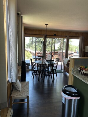 A dining area with a table and chairs, a trash bin, and a view of the outdoors through large windows.