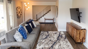 A living room with a grey sofa, a wooden coffee table, a patterned rug, a staircase, and a TV mounted on the wall. - Eye-catching Home and Yard w/ Luxurious Hot Tub, Patio, and Private Trail Access (Port Angeles)