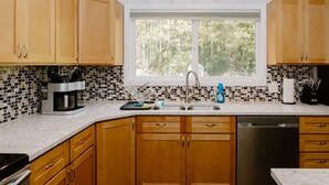 A kitchen with wooden cabinets, a coffee maker, a sink, and a window with a view of trees. - Eye-catching Home and Yard w/ Luxurious Hot Tub, Patio, and Private Trail Access (Port Angeles)