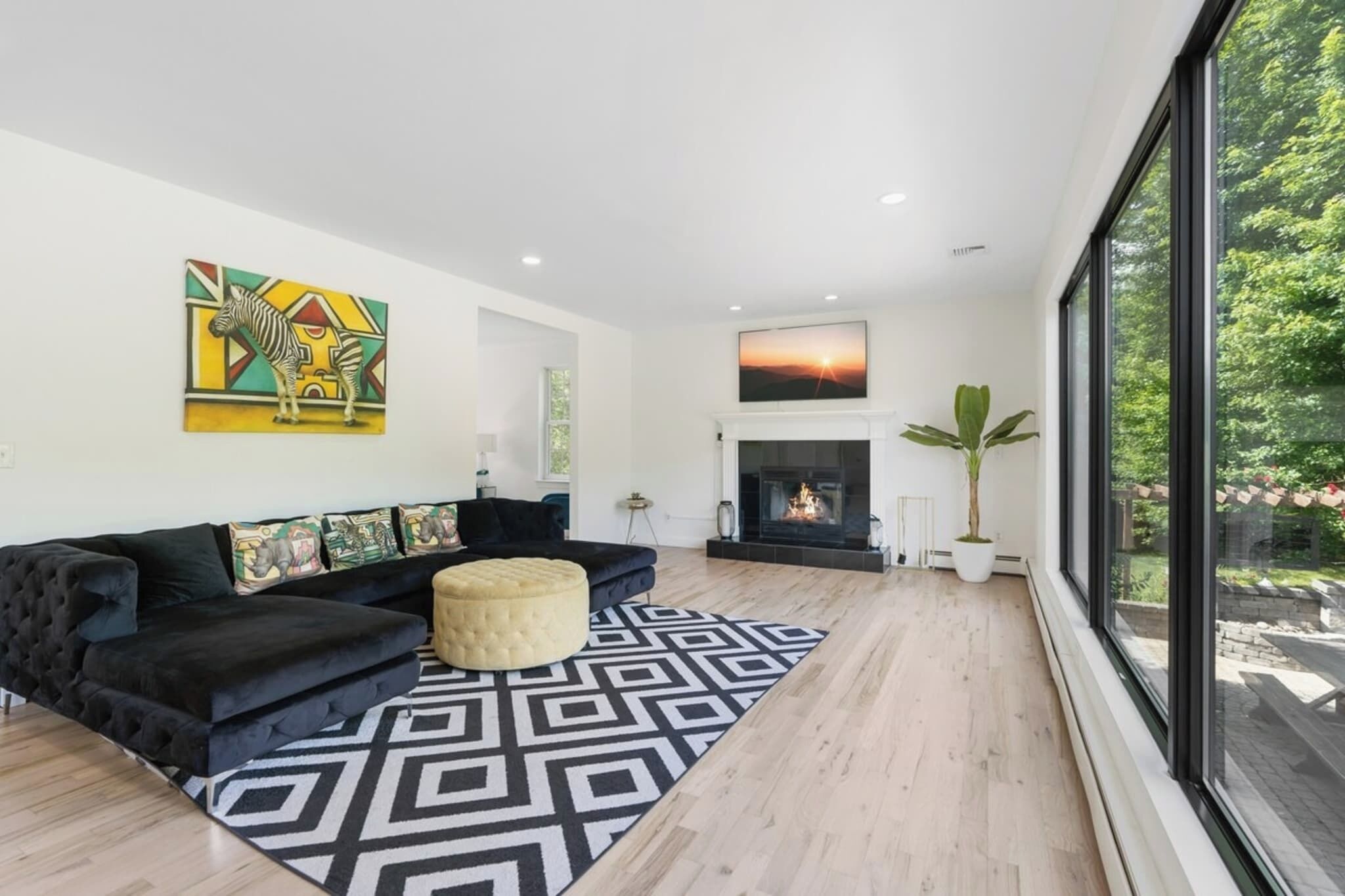 A modern living room with a black sofa, a patterned rug, a fireplace, and a large window with a view of greenery.
