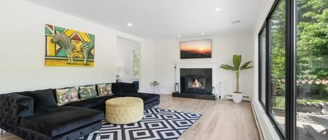A modern living room with a black sofa, a patterned rug, a fireplace, and a large window with a view of greenery.