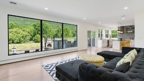 A modern living room with a large window, a sofa, ottoman, and a kitchen in the background.