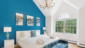 A bedroom with a bed, two bedside tables, a bench, a chandelier, and a large window.
