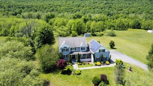 A large two-story house with a solar panel on the roof, surrounded by lush greenery and a hilly landscape.