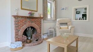 Een woonkamer met een haard van baksteen, een ronde tafel, een comfortabele fauteuil, een spiegel en een raam met gordijnen.