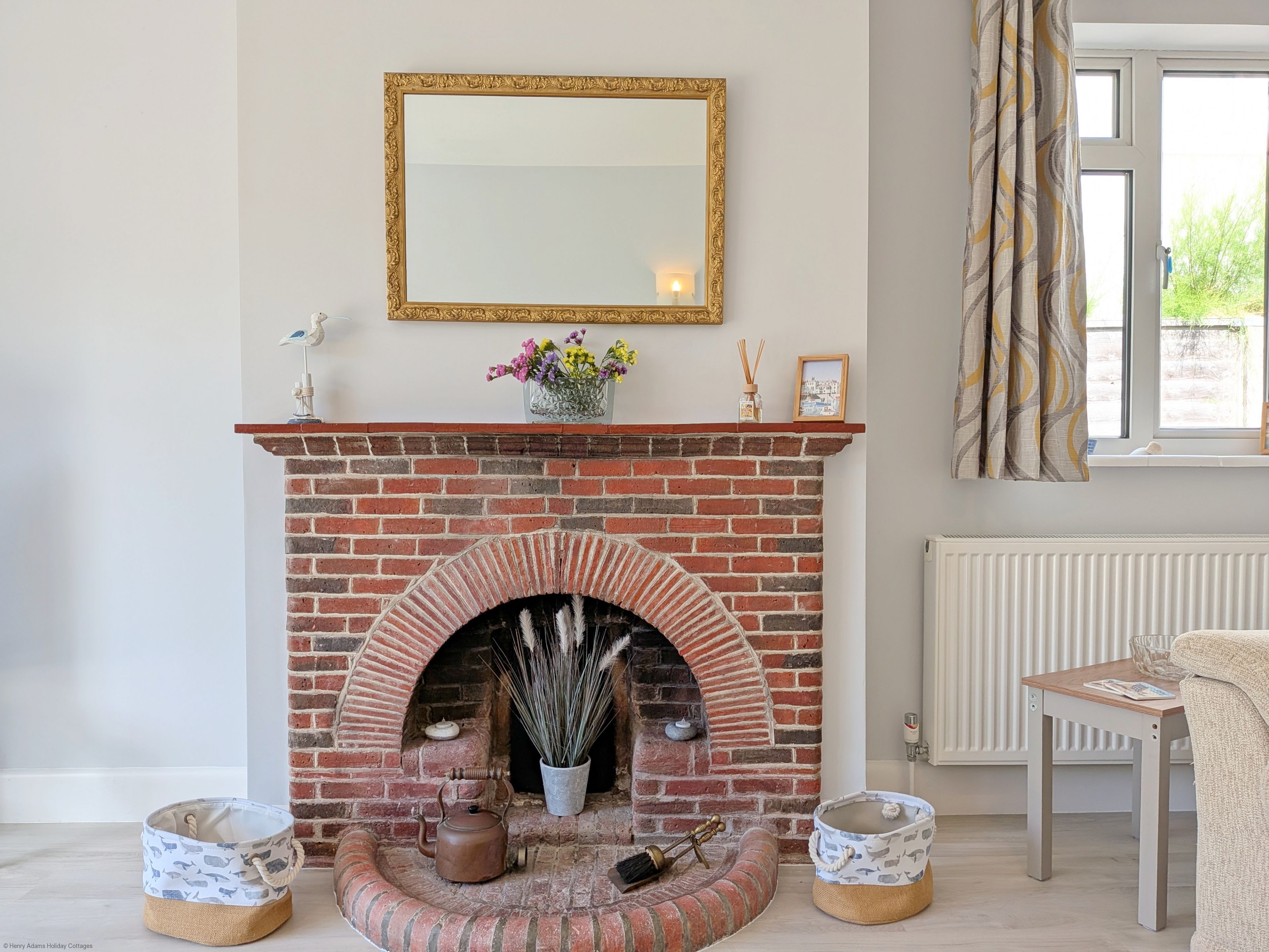 A room with a brick fireplace, a mirror, a window with curtains, and a table with a magazine.