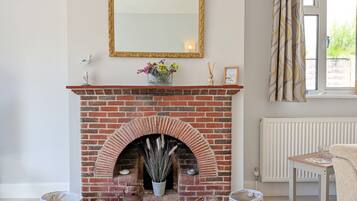 A room with a brick fireplace, a mirror, a window with curtains, and a table with a magazine.
