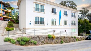 A two-story building with a white facade, multiple windows, and a stone retaining wall with plants.