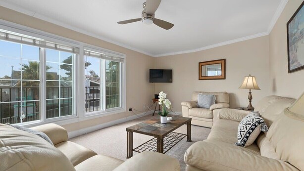 A living room with a ceiling fan, a flat-screen TV, a beige sofa, a coffee table, and large windows.