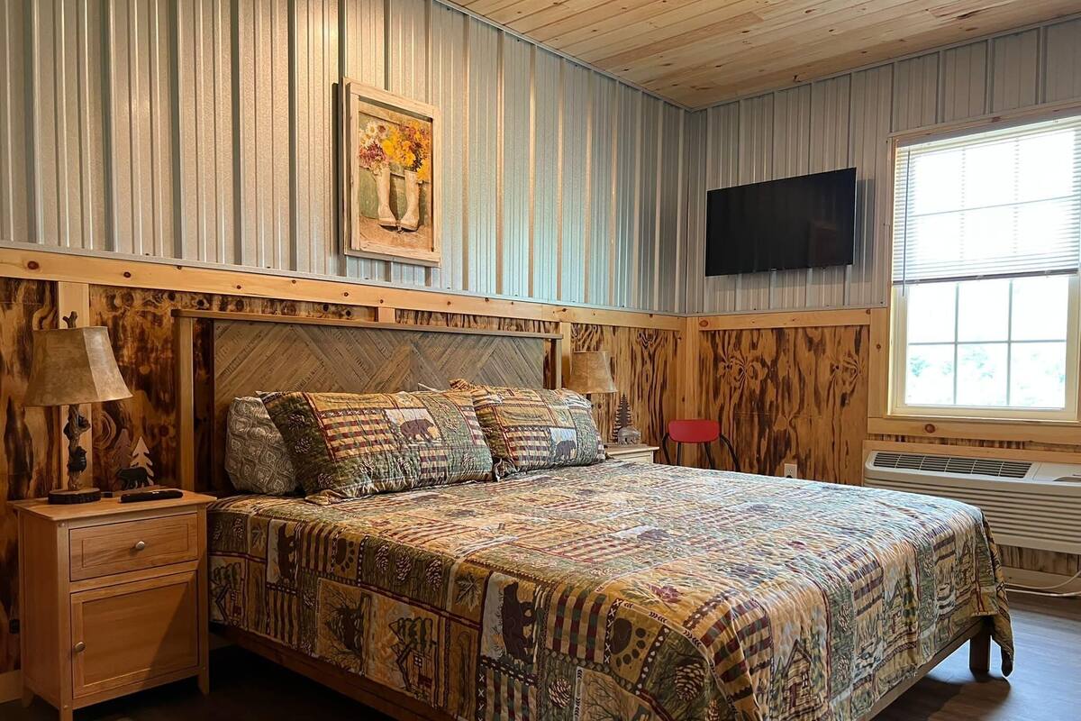 Une chambre à coucher avec une tête de lit en bois, un lit avec une literie à motifs, une table de chevet avec une lampe, un fauteuil rouge et un téléviseur fixé au mur.