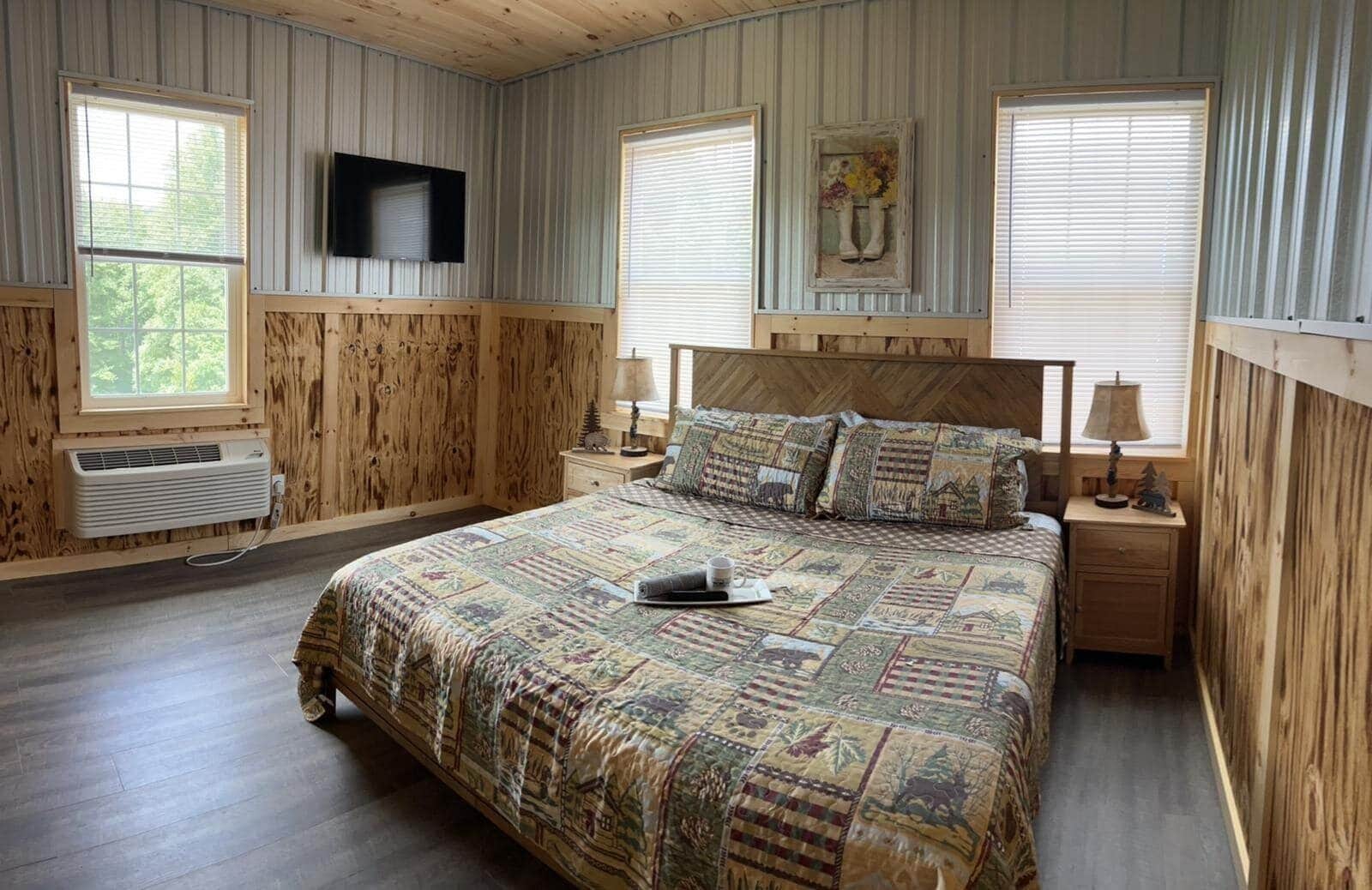 A bedroom with a large bed, wooden walls, a mounted TV, and a window with blinds.