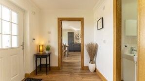 Un pasillo con suelo de madera, una puerta a la izquierda, una mesita con una lámpara y una planta, y un jarrón con ramas a la derecha.