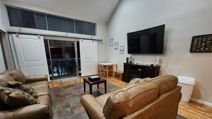 A living room with a brown leather sofa, a television mounted on the wall, and a sliding glass door leading to a balcony. - Relaxing Lake Conroe Condo overlooking the pool and lake (Montgomery)