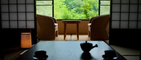 Una habitación japonesa tradicional con suelo de tatami, una mesa baja con tetera y tazas, y vistas a un paisaje verde exuberante a través de amplias ventanas.