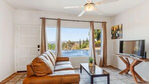 A living room with a leather sofa, a coffee table, a TV on a wooden stand, and a view of a pool and landscape through large windows.
