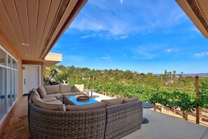 A covered outdoor seating area with a wicker sofa and a table, overlooking a vineyard.
