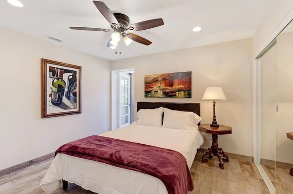 A bedroom with a bed, a ceiling fan, a painting, a lamp, and a door.