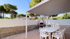 En udendørs patio med en stenvæg, en hvid pergola, et rundt bord med hvide stole og en indbygget pizzaovn.
