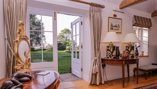 A room with a wooden table, a lamp, a rug, and a view of a garden through a door. - Little Pontus (Canterbury)