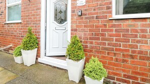 Uma casa de tijolos com porta branca, uma janela pequena e uma placa com a inscrição "Cedar Cottage". Há três vasos com plantas em frente à porta.