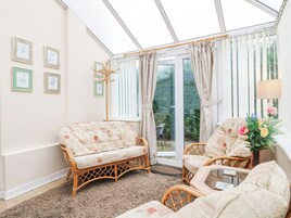 A conservatory with wicker furniture, framed artwork on the walls, and a view of greenery outside.