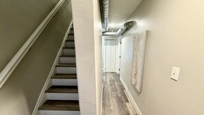 A hallway with a wooden staircase, a door at the end, and a wall-mounted light fixture.