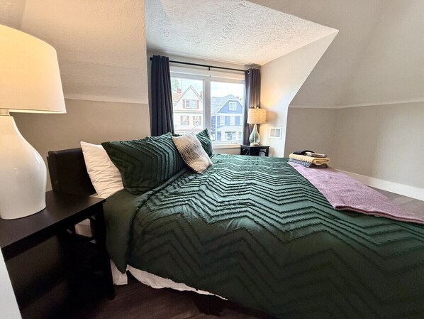 Ein Schlafzimmer mit Bett, Nachttisch, Lampe, Fenster mit Vorhängen und Blick auf Gebäude.