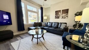 A living room with a glass coffee table, a dark leather sofa, a TV on the wall, and a large window with curtains.