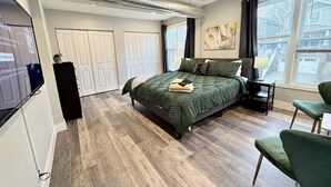 A bedroom with a large bed, a desk, and a chair. There are two doors and a window with curtains.