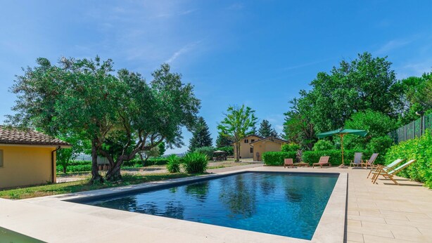 Une piscine rectangulaire, bordée d’une terrasse carrelée, entourée de verdure et de quelques bâtiments.