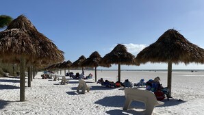 Ein Strand mit Reetsonnenstühlen und Plastikstühlen, ein klarer Himmel und Fußabdrücke im Sand.