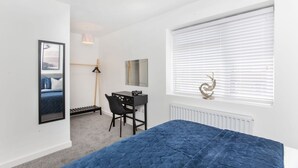 Et soveværelse med seng, skrivebord, stol, radiator, et vindue med persienner og et spejl på væggen.