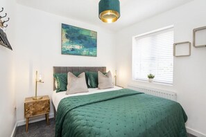 A bedroom with a bed, a bedside table, a lamp, a window with blinds, and a painting on the wall.