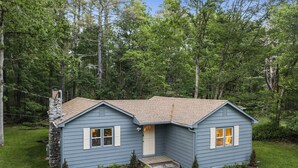A small, single-story house with a stone chimney, a front porch, and a pathway leading to the entrance. - Retro Countryside Cabin | Cozy Upstate NY Private Retreat! (Eldred)